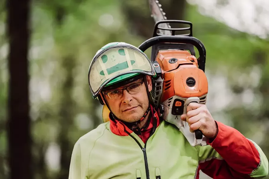 Ein Mann mit Schutzkleidung und Brille trägt eine orangefarbene Kettensäge auf seiner Schulter in einem Wald.