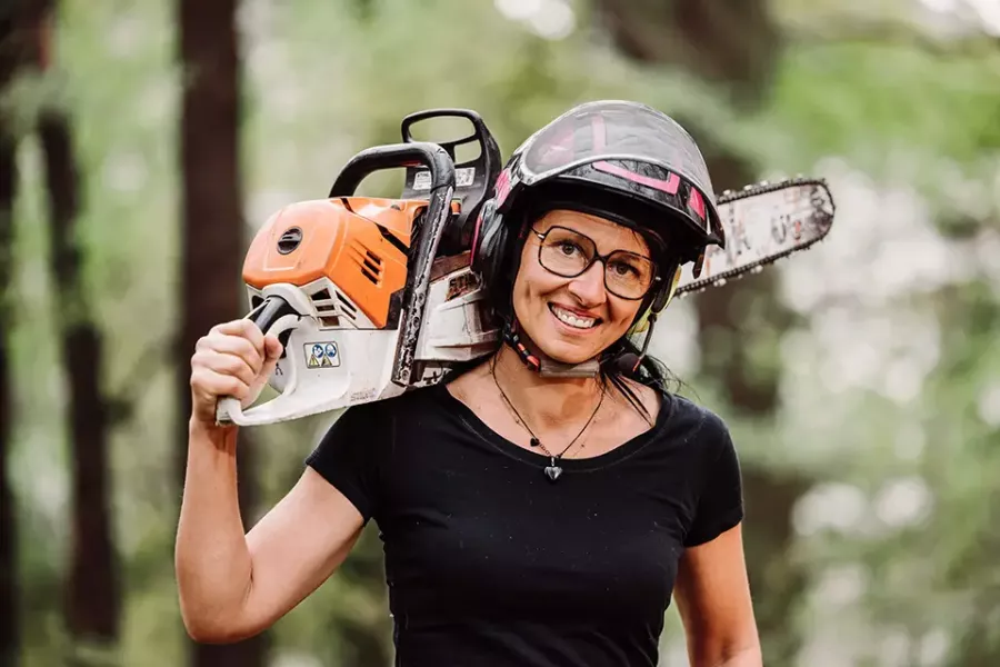 Eine lächelnde Frau mit Brille und Helm hält eine Kettensäge auf ihrer Schulter, während sie im Freien in einem Waldgebiet steht.