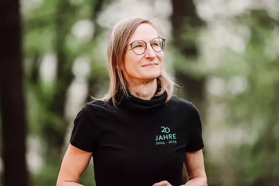 Eine Frau mit Brille und schulterlangem blondem Haar steht im Freien und trägt ein schwarzes T-Shirt mit der Aufschrift 20 Jahre 2004-2024. Sie blickt leicht nach oben und zur Seite, wobei die Bäume im Hintergrund verschwimmen.