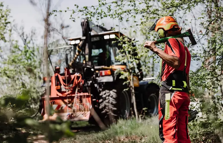Eine Person in roter Schutzkleidung und mit Helm trägt eine Axt über der Schulter, während sie in einem Waldgebiet in der Nähe schwerer Maschinen steht, möglicherweise bei Holzfäller- oder Forstarbeiten.