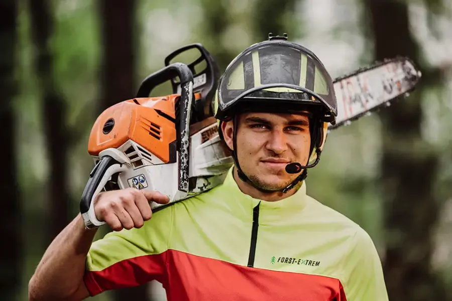 Ein Mann, der einen Helm und ein Warnschutzhemd trägt, stützt eine Kettensäge auf seine Schulter. Er steht im Freien in einem Waldgebiet und blickt mit entschlossener Miene nach vorne.