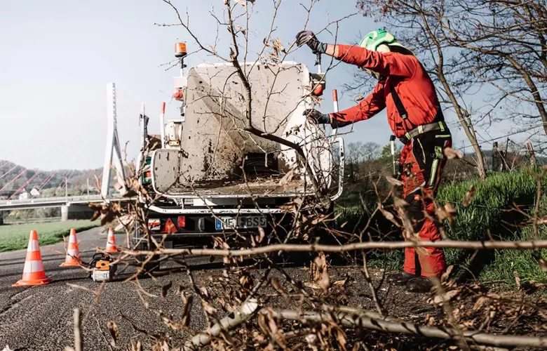 Ein Arbeiter in Schutzkleidung führt auf einer mit Verkehrskegeln gesäumten Straße Äste in eine Holzhackmaschine ein. Baumreste liegen auf dem Boden verstreut, und im Hintergrund ist eine Brücke zu sehen.