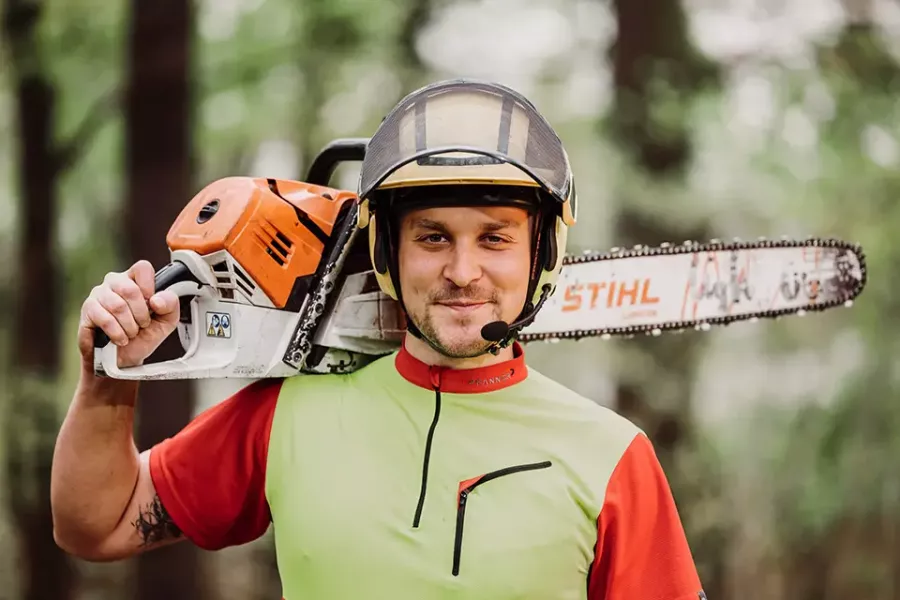 Ein lächelnder Mann mit Schutzausrüstung hält eine Kettensäge auf seiner Schulter in einer Waldlandschaft. Er ist mit einem grün-roten Arbeitshemd und einem Schutzhelm bekleidet.