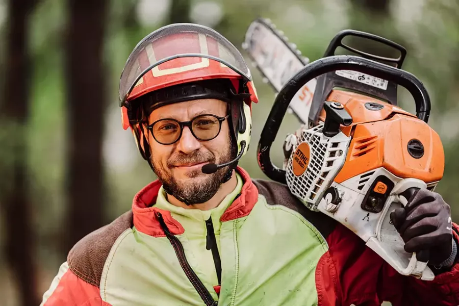 Ein Mann, der eine Sicherheitsausrüstung trägt, darunter einen Helm, eine Brille, Handschuhe und ein Headset, lächelt, während er in einem Waldstück eine Kettensäge über der Schulter hält.